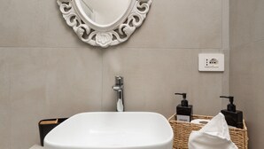 Et badeværelse med en hvid vask, et spejl med detaljeret udsmykning, en vandhane og toiletartikler i en fletkurv.