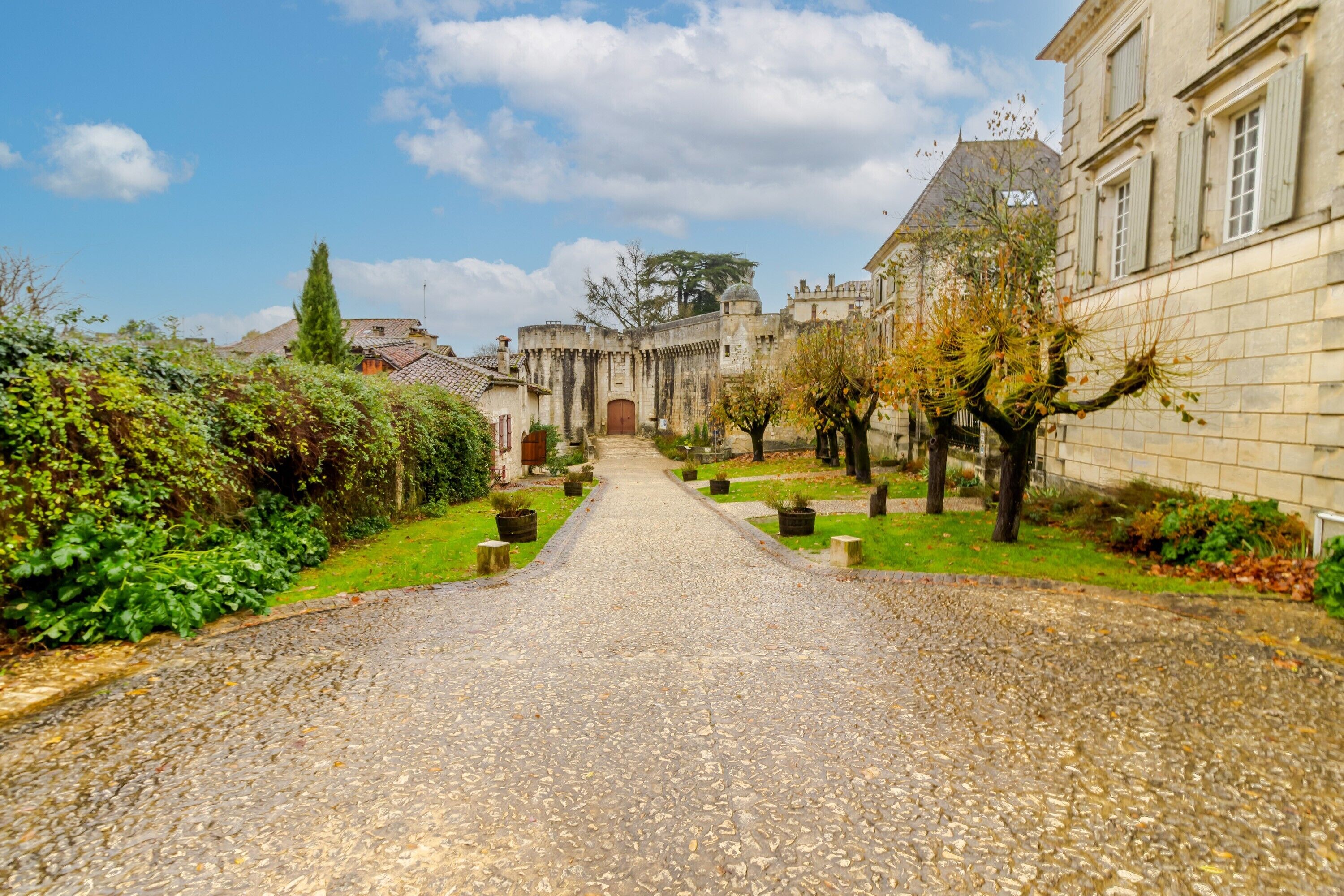 A cobblestone pathway leading to a historic building with a central entrance, surrounded by greenery and trees.