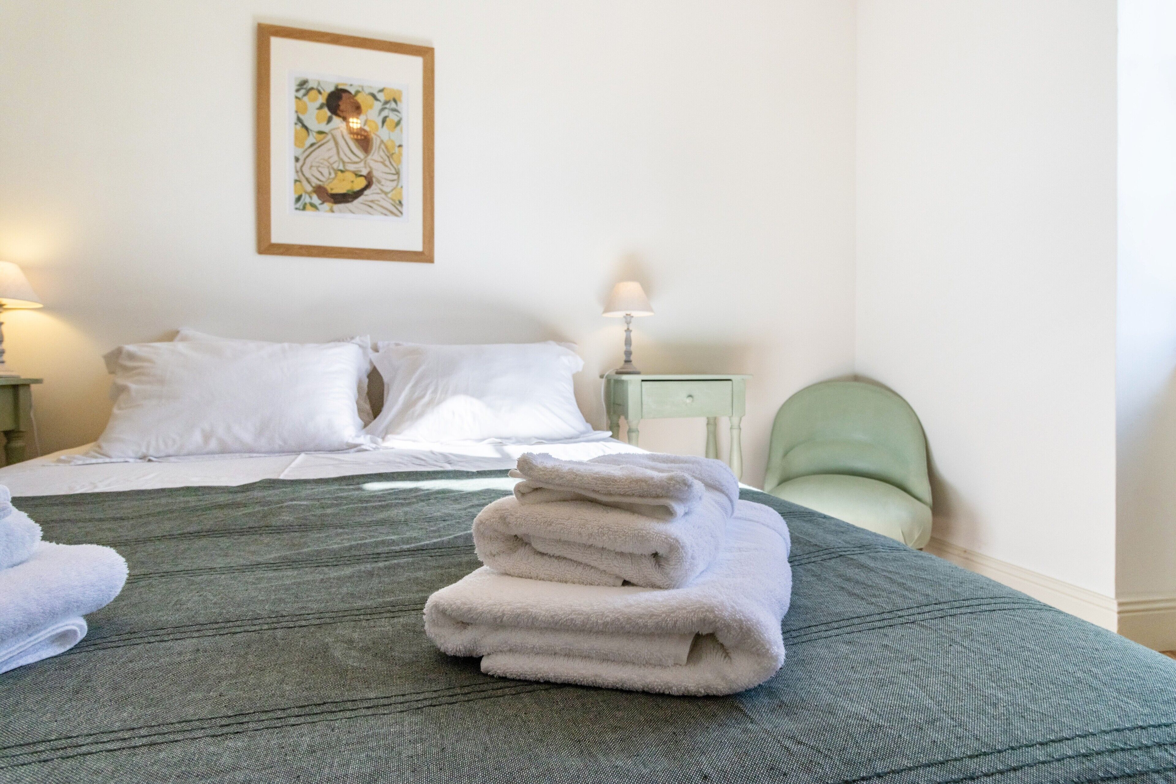 A neatly made bed with white linens, a green textured bedspread, and a stack of folded white towels on the bed.