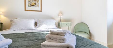 A neatly made bed with white linens, a green textured bedspread, and a stack of folded white towels on the bed.