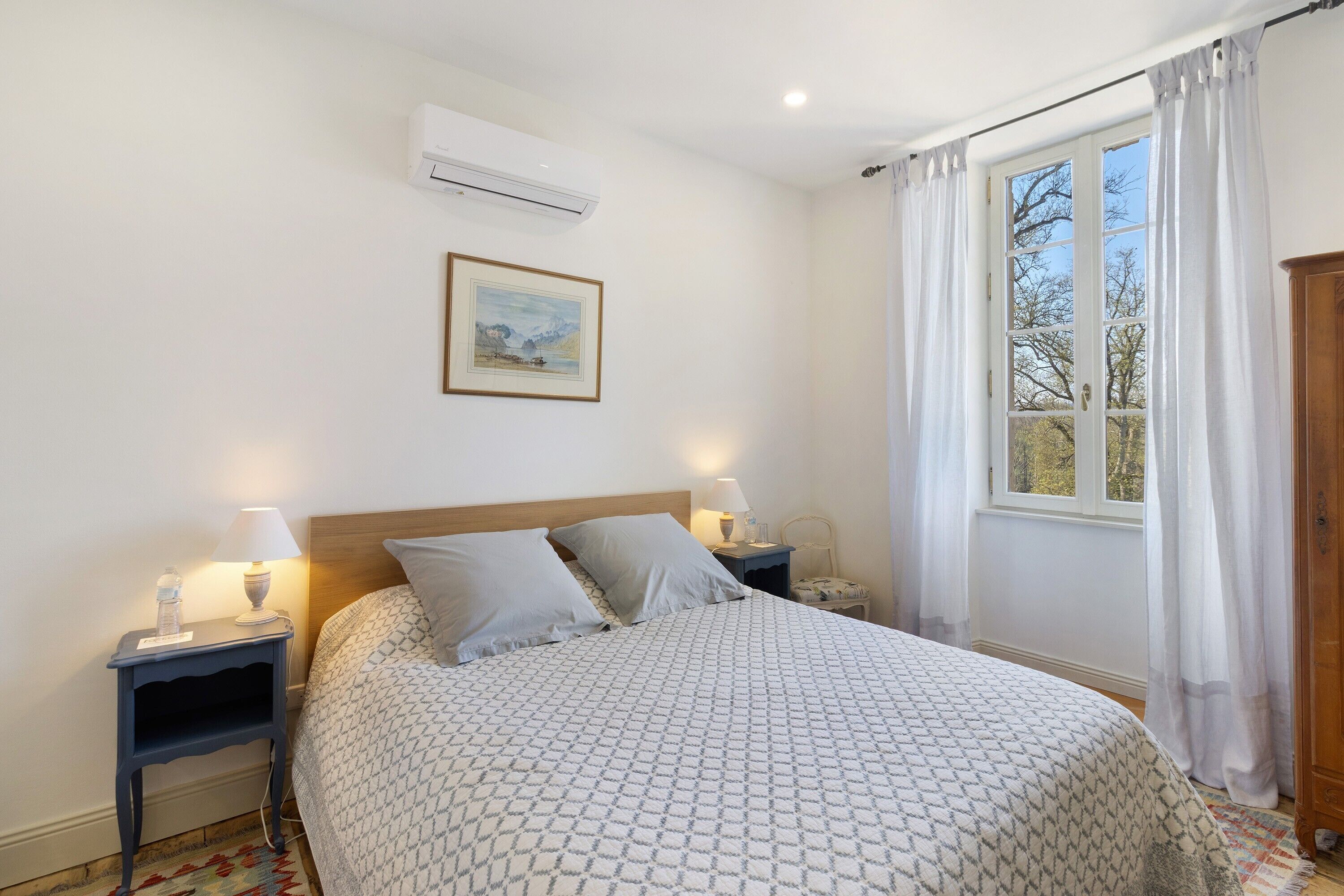 Un dormitorio con cama, dos mesitas de noche con lámparas, una mesita auxiliar, una ventana con cortinas, un equipo de aire acondicionado y un cuadro en la pared.