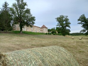 A stone building with a red roof, surrounded by trees and hay bales in a grassy field.