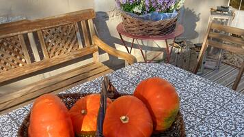 A wicker basket with pumpkins on a patterned tablecloth, a wooden bench, a small round table with a wicker basket, and a potted plant with purple flowers.