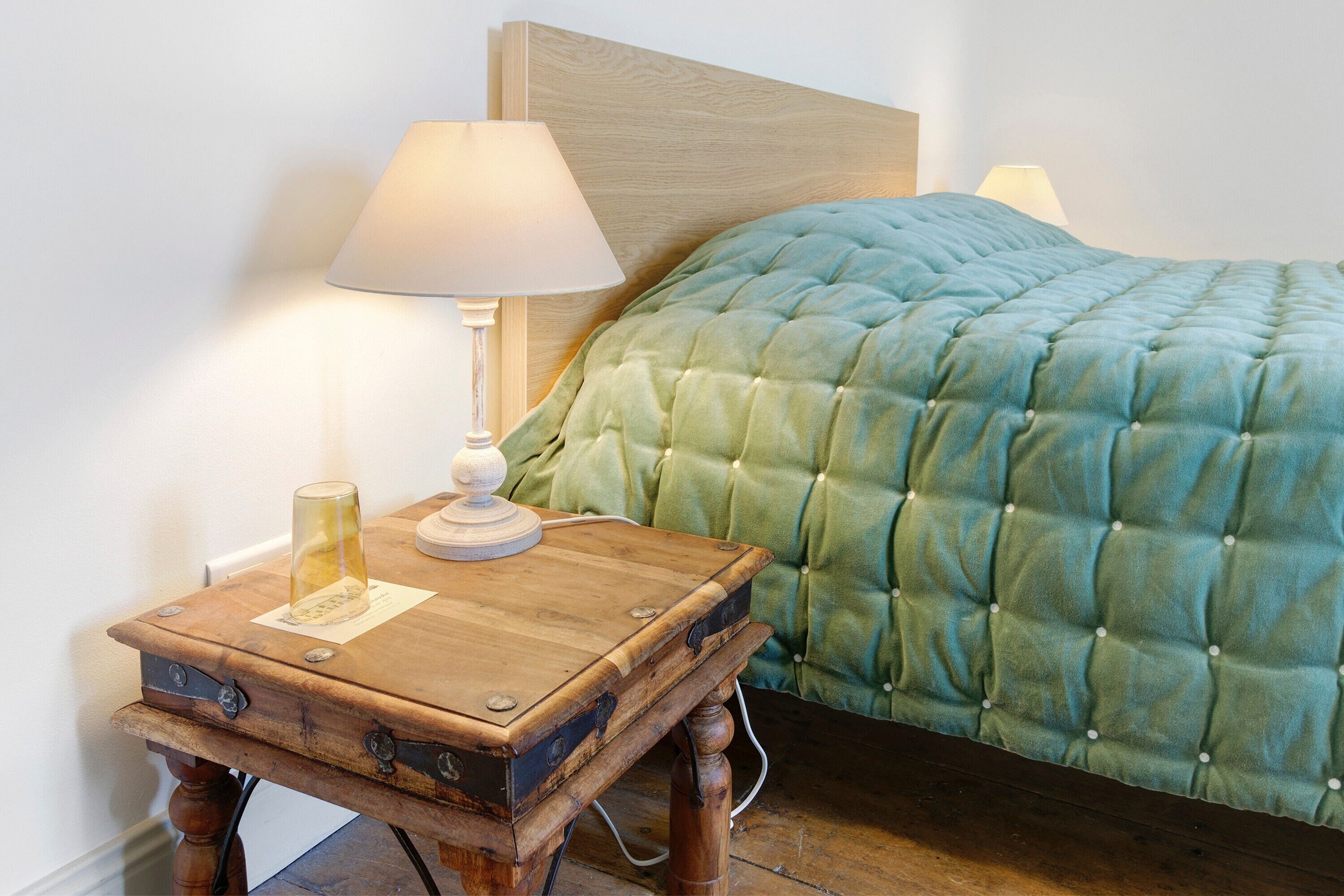 A bed with a green quilt, a wooden nightstand with a lamp, and a glass on the nightstand.