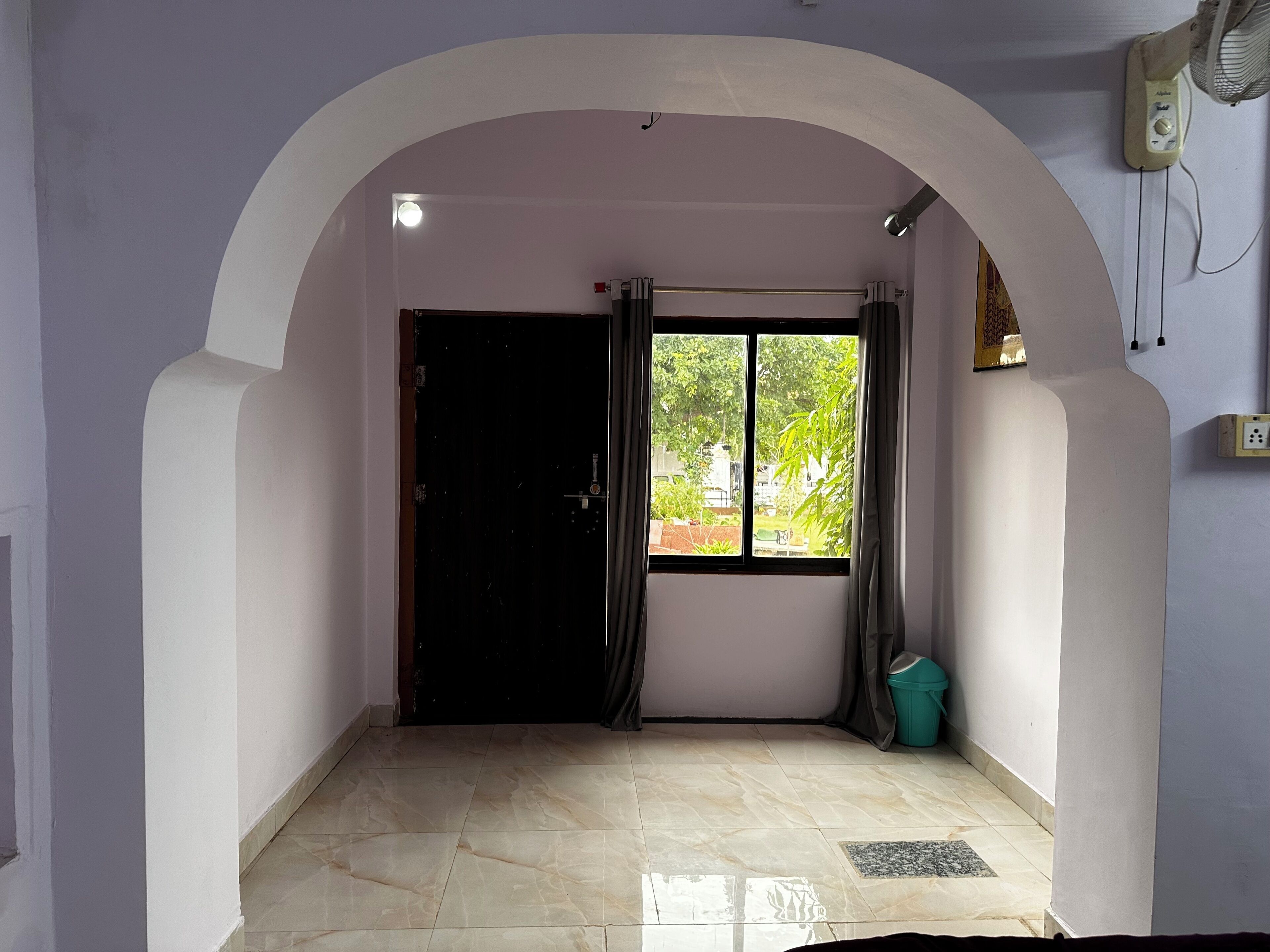 Una habitación con suelo de baldosas, una puerta negra, un ventanal con cortinas y un cubo de basura pequeño.