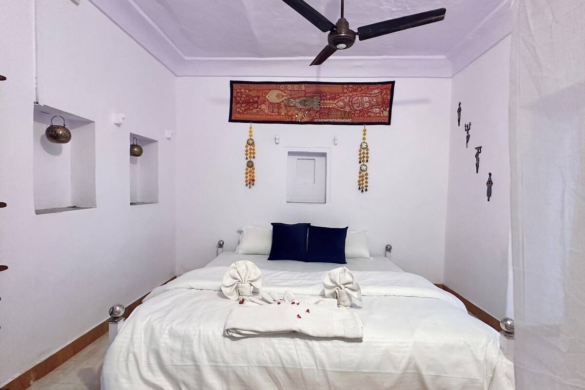 Een netjes ingerichte slaapkamer met een bed, kussens, een plafondventilator en een decoratief wandtapijt.