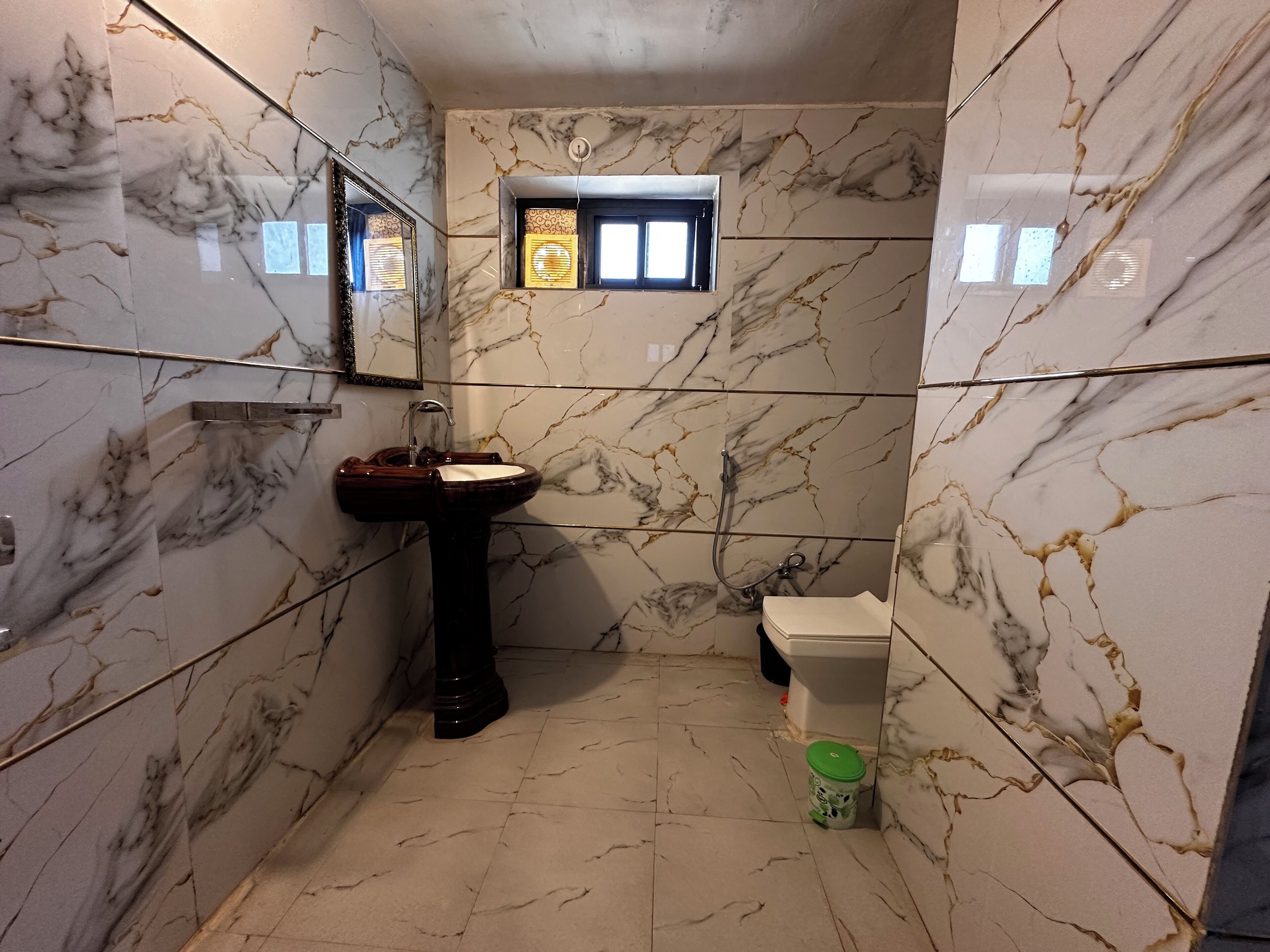 A bathroom with marble walls, a pedestal sink, a window, and a toilet.