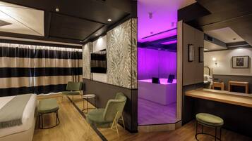Een moderne hotelkamer met een bed, groene stoelen, een klein tafeltje en een receptiegedeelte met paars licht.