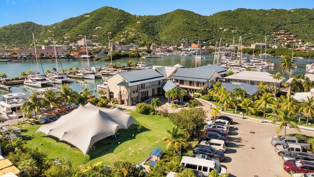 Un puerto deportivo con barcos, un edificio grande con techo inclinado y un estacionamiento con coches.