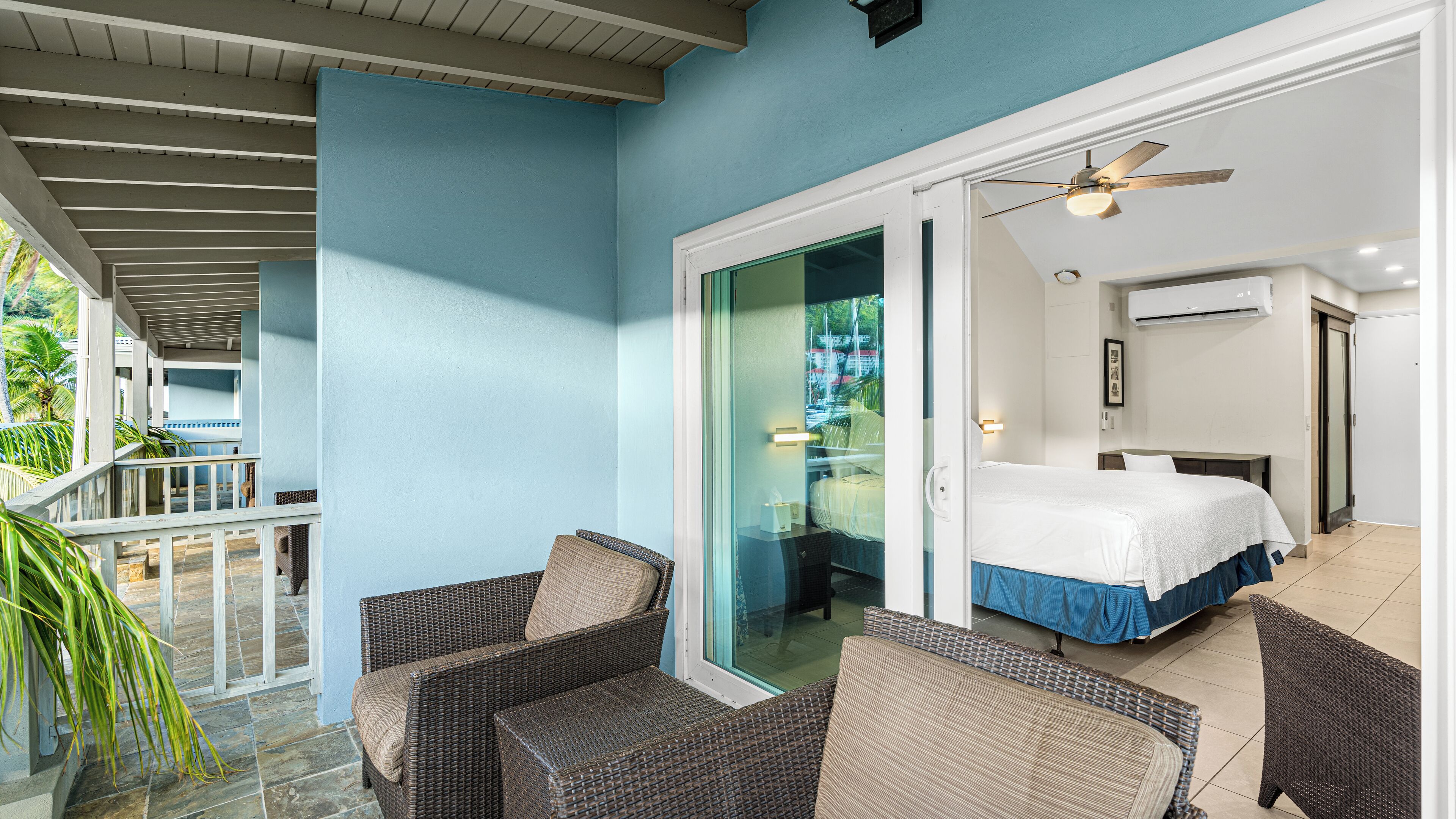Une chambre d’hôtel avec un balcon, des meubles en osier, un lit avec une couvre-lit bleue et un ventilateur de plafond.