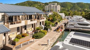 Un puerto deportivo con varios yates amarrados, un edificio con balcones que dan al agua, y un paisaje montañoso al fondo.