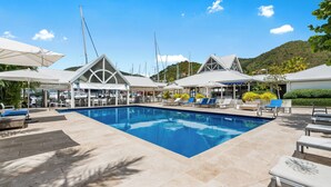 Un área de piscina con sillones de descanso y sombrillas, rodeada de zonas de estar cubiertas y barcos al fondo.