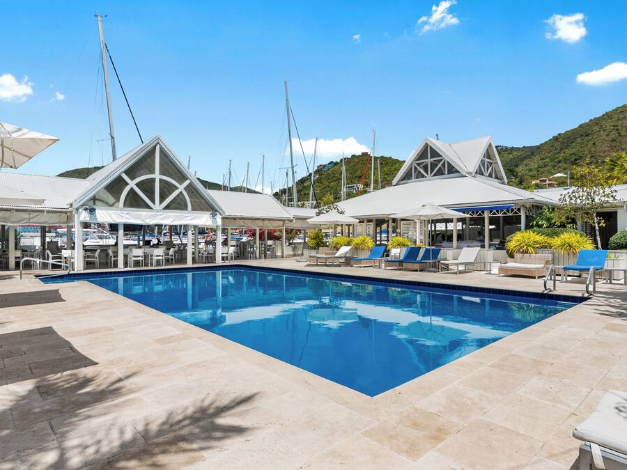 A pool area with lounge chairs and umbrellas, surrounded by covered seating areas and boats in the background.