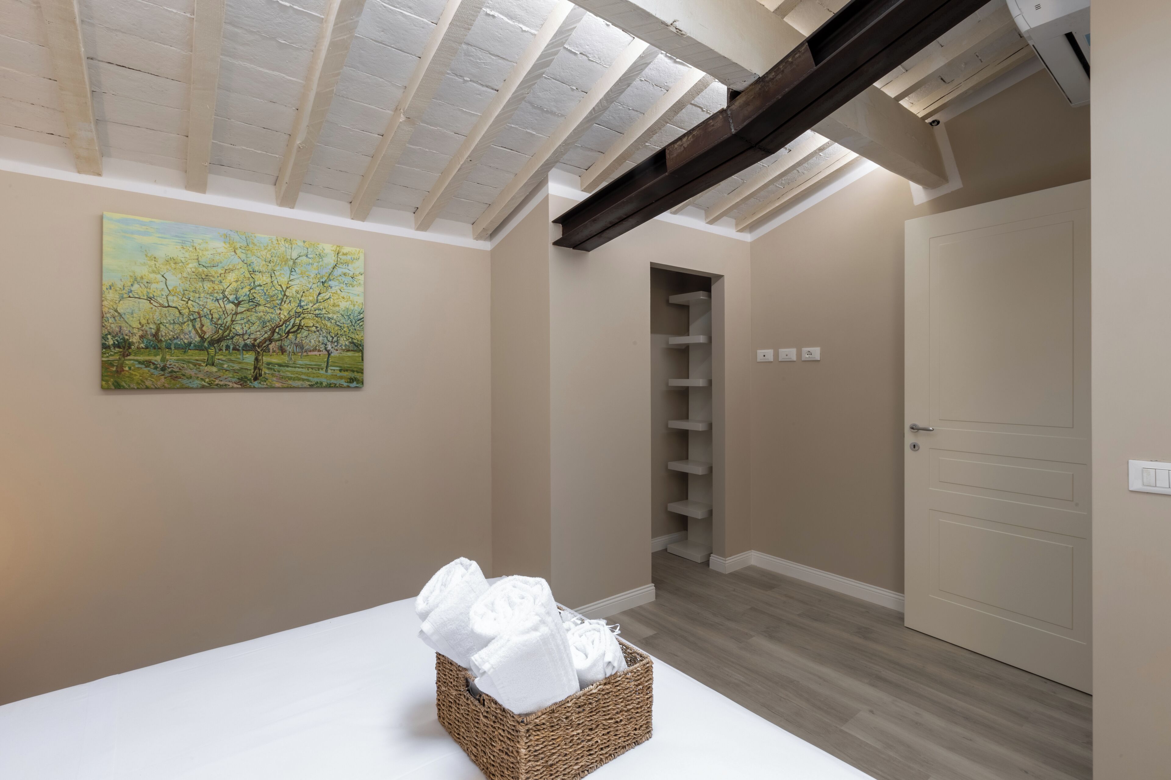 A room with a high ceiling, a painting of trees, a wicker basket with towels, a door, and a built-in shelf.
