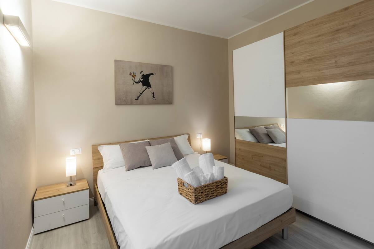 Une chambre à coucher bien rangée, avec un lit, une lampe de chevet, une table de nuit, une œuvre d’art accrochée au mur et un miroir.