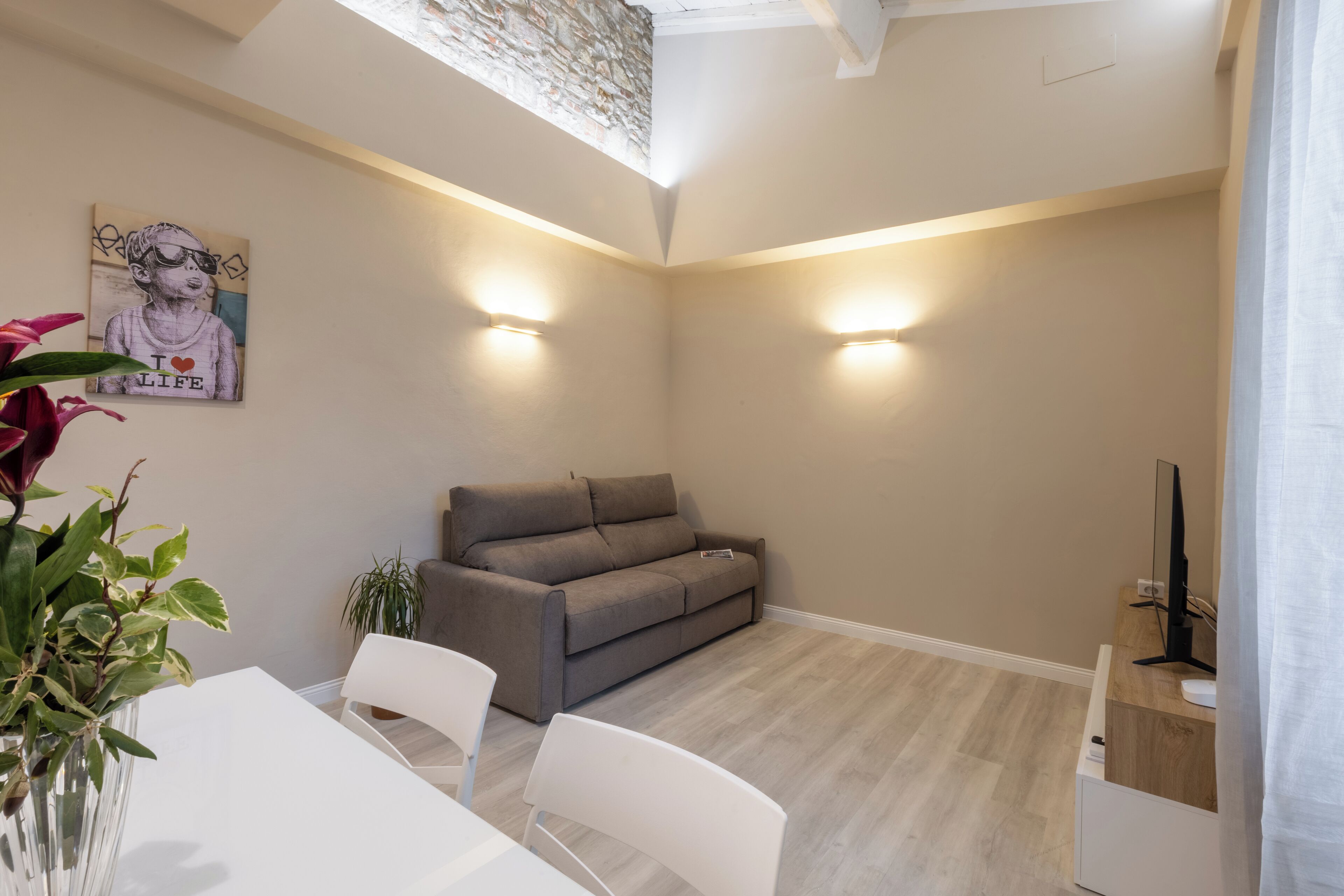 A modern living room with a grey sofa, a white dining table, and a TV on a wooden stand.