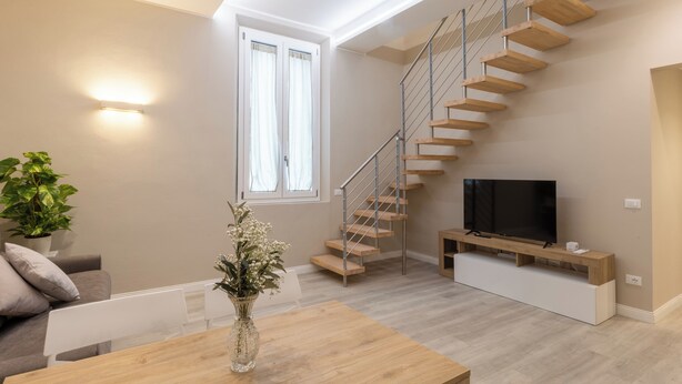 A modern living room with a wooden staircase, a flat-screen TV, and a dining area with a vase of flowers. - Orsini 98 Apartments by Mamo Florence (Florence)