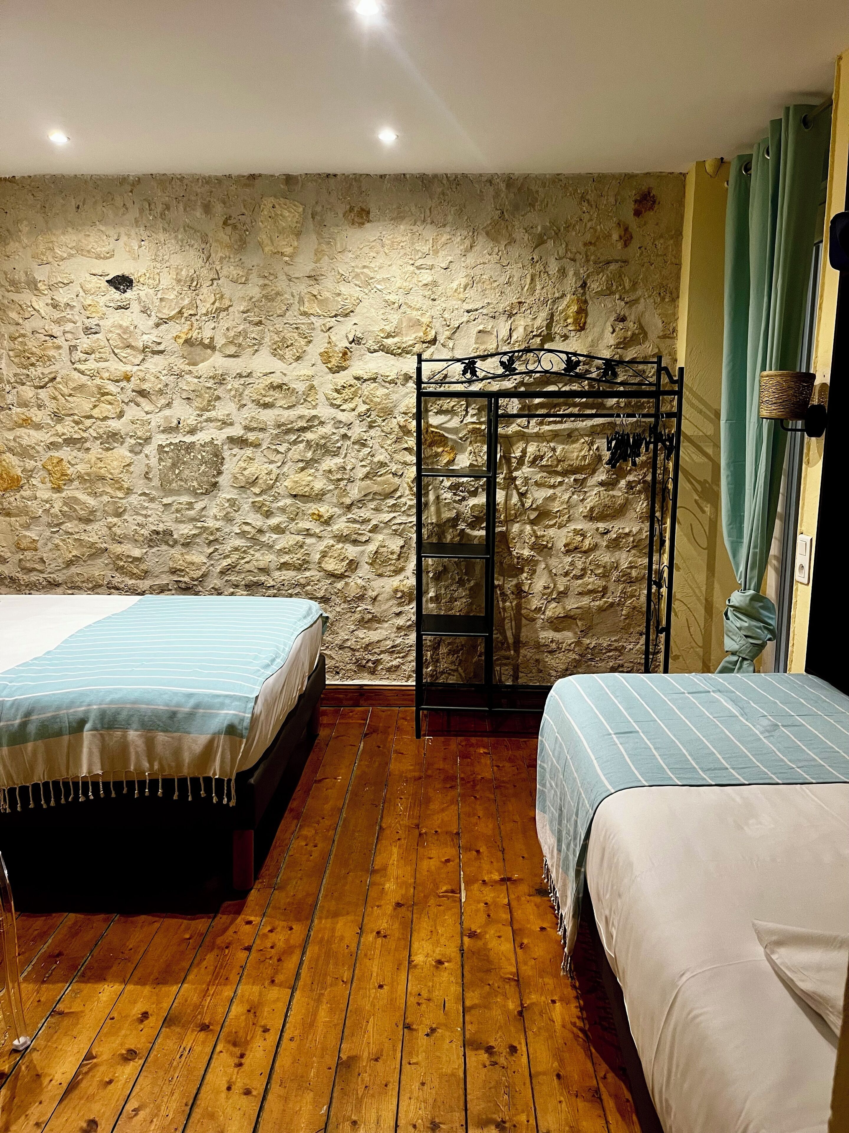 Une chambre avec deux lits, un plancher en bois, des murs en pierre, un étagère en métal et une fenêtre avec des rideaux.