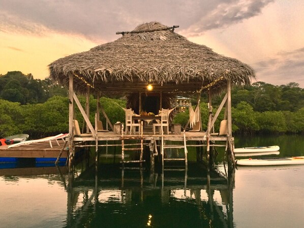 A thatched-roof wooden structure with a table and chairs, surrounded by calm water and greenery.