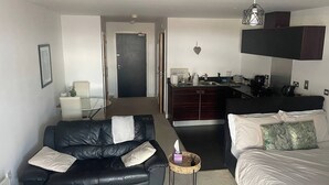 Een compacte hotelkamer met een zwarte leren bank, een bed met witte beddengoed, een klein keukenhoekje en een eettafel.