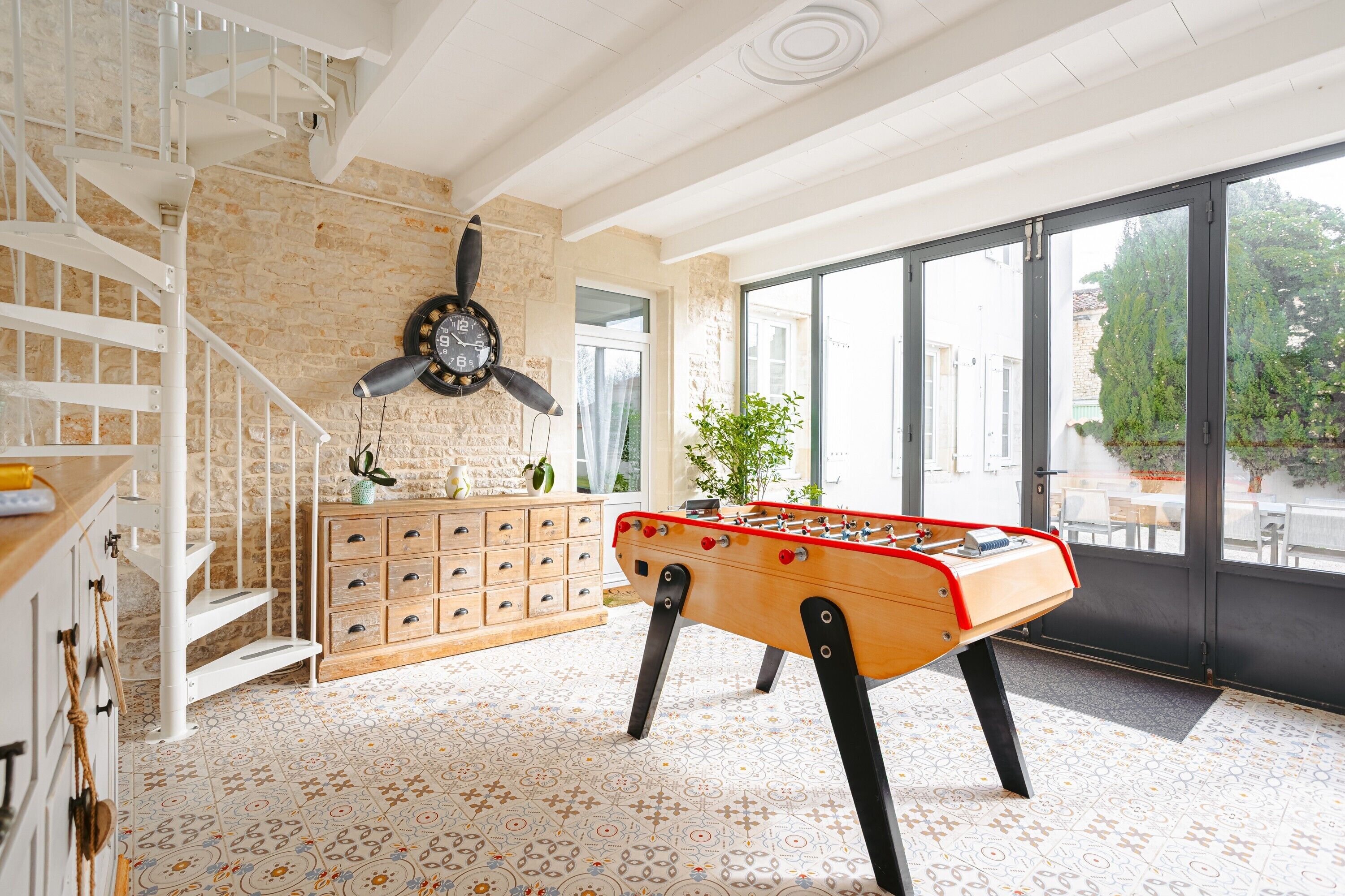Une pièce avec une table de baby-foot, une horloge, un escalier et une grande fenêtre.