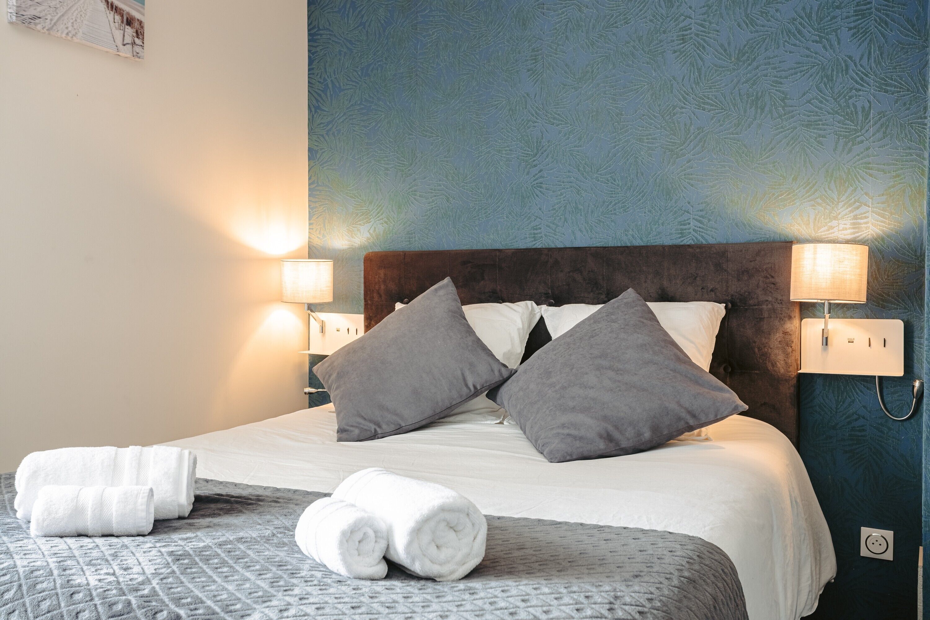 A neatly made bed with pillows, a bedside lamp, and rolled towels on a patterned bedspread.