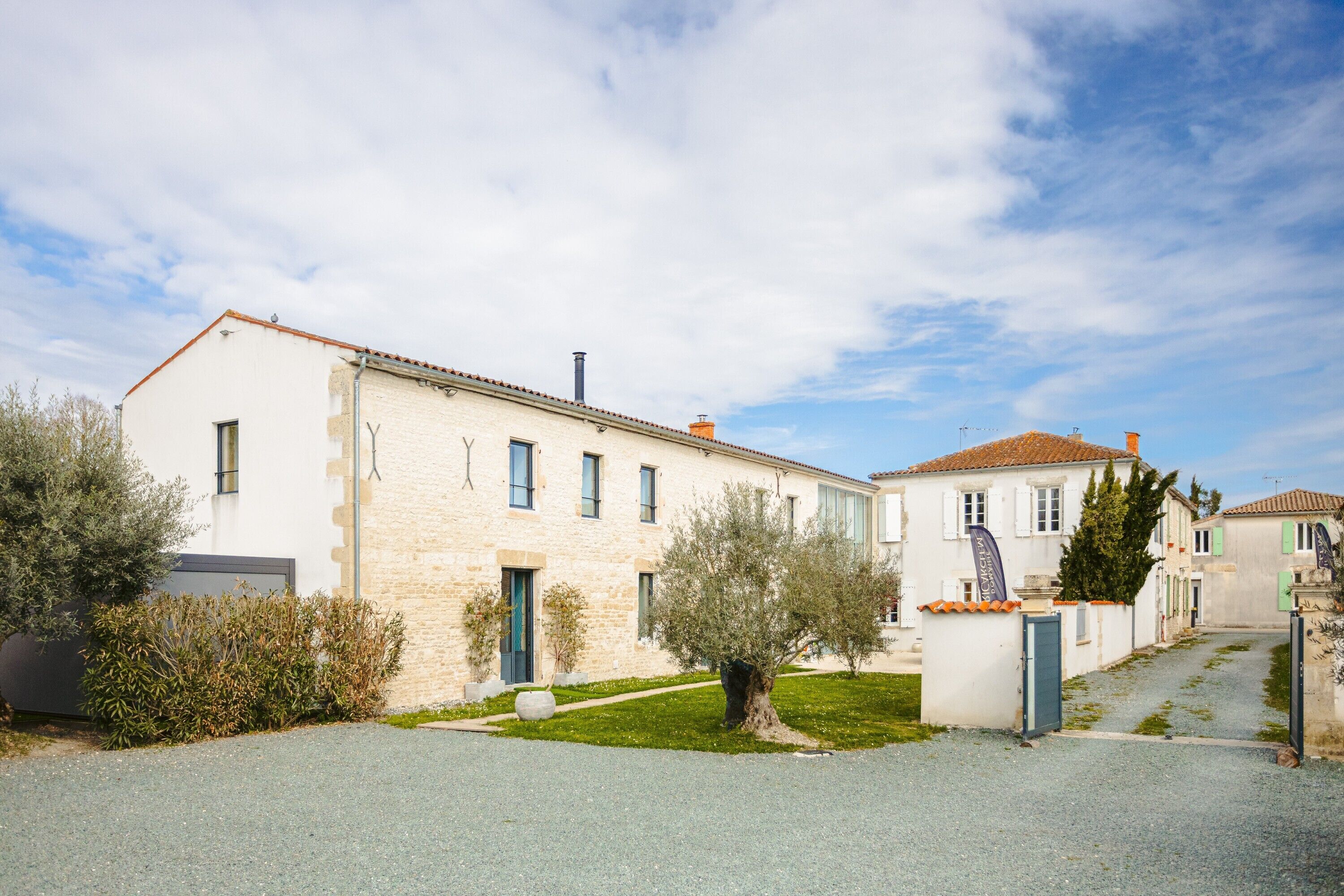 Un bâtiment à deux étages avec une façade en pierre, une allée en gravier et un petit arbre dans le jardin.