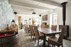 Een eetgedeelte met een lange tafel, rieten stoelen, een houten vat en een zwarte kachel.