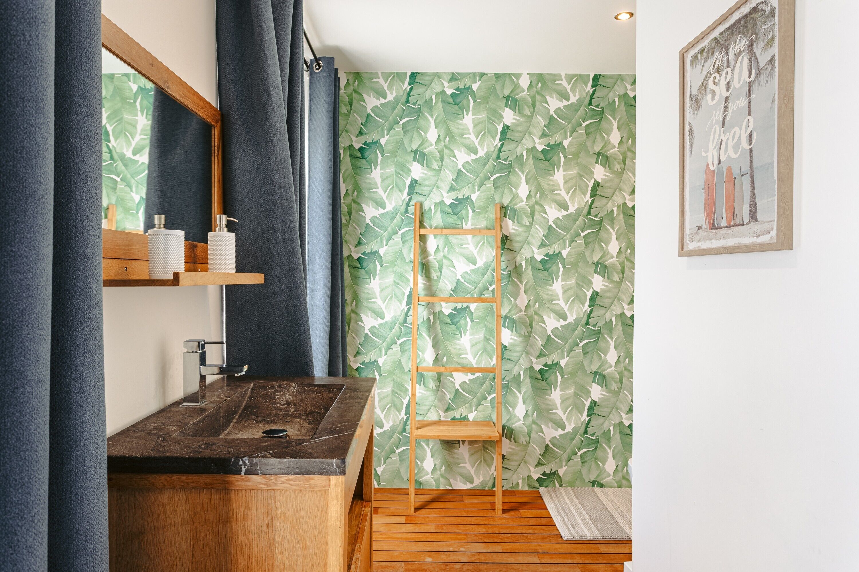 Une salle de bain avec un meuble-lavabo en bois, un miroir, une étagère avec des articles de toilette, une échelle en bois et un papier peint à motifs de feuilles tropicales.
