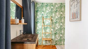 Une salle de bain avec un meuble-lavabo en bois, un miroir, une étagère avec des articles de toilette, une échelle en bois et un papier peint à motifs de feuilles tropicales.