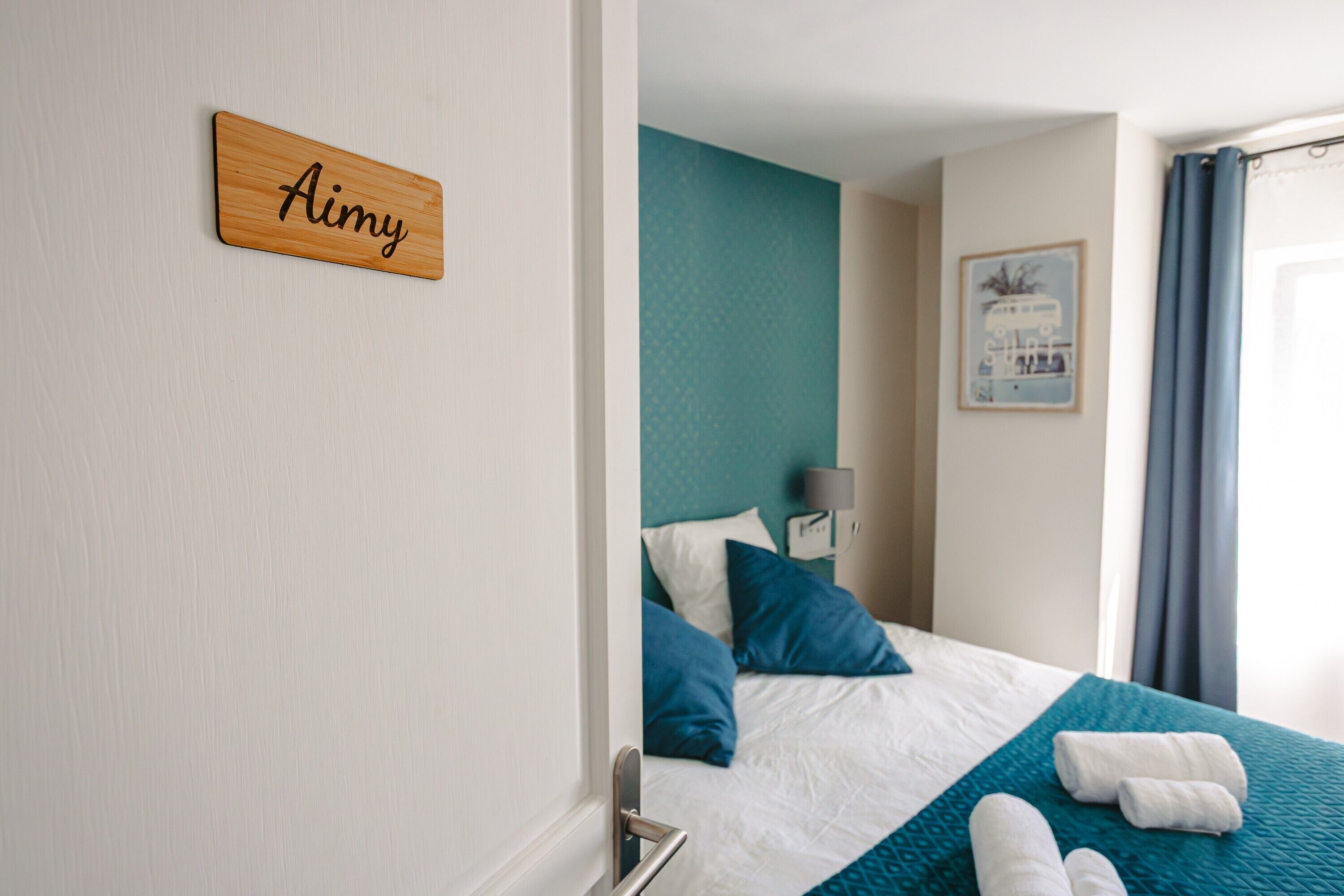 Una cama bien hecha con una colcha color turquesa, almohadas blancas y un letrero de madera con el nombre 'Aimy' en la pared.