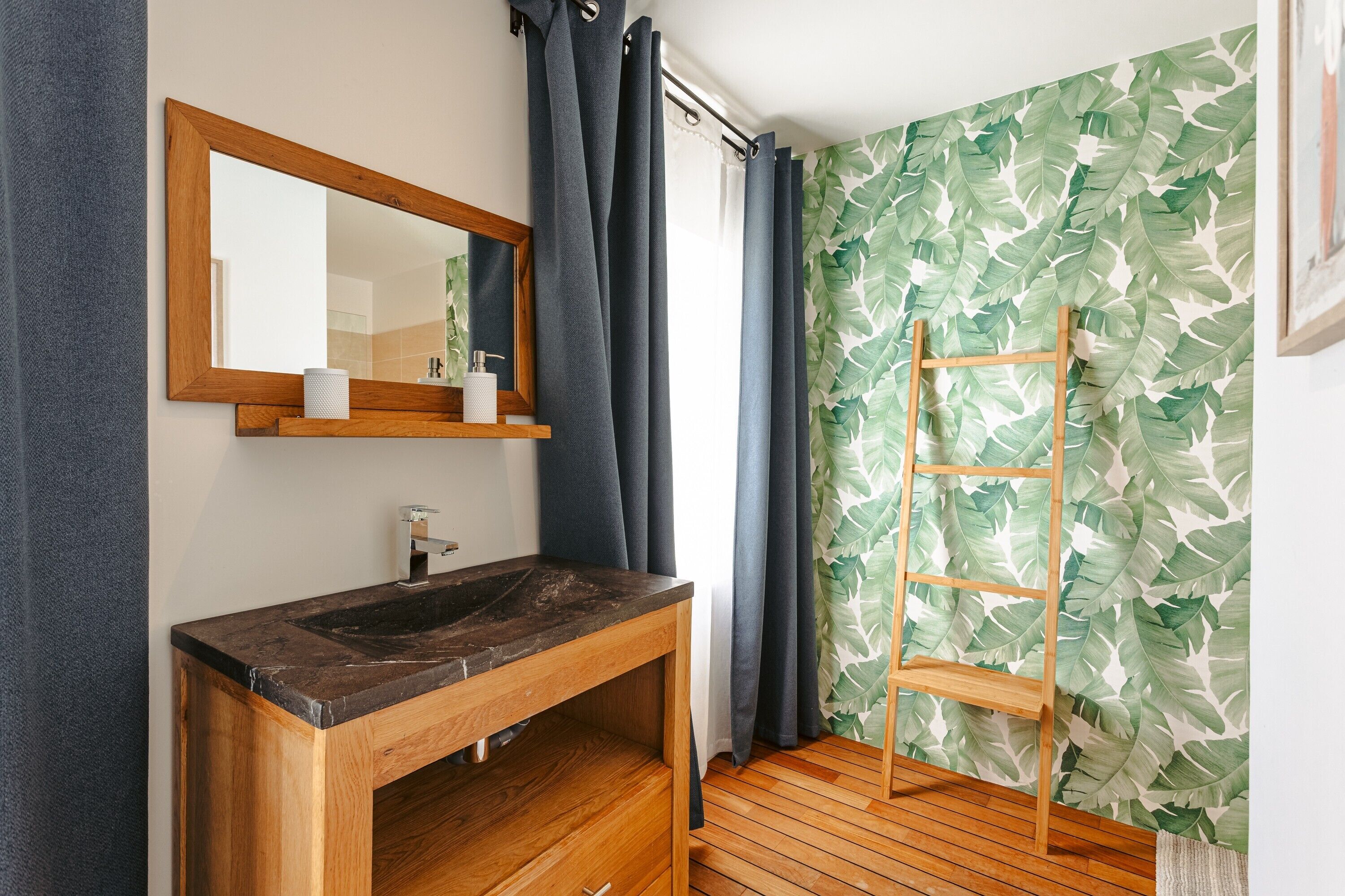 A bathroom with a wooden vanity, a mirror, a shelf with toiletries, a shower curtain, and a wooden ladder against a wall with a leaf-pattern wallpaper.