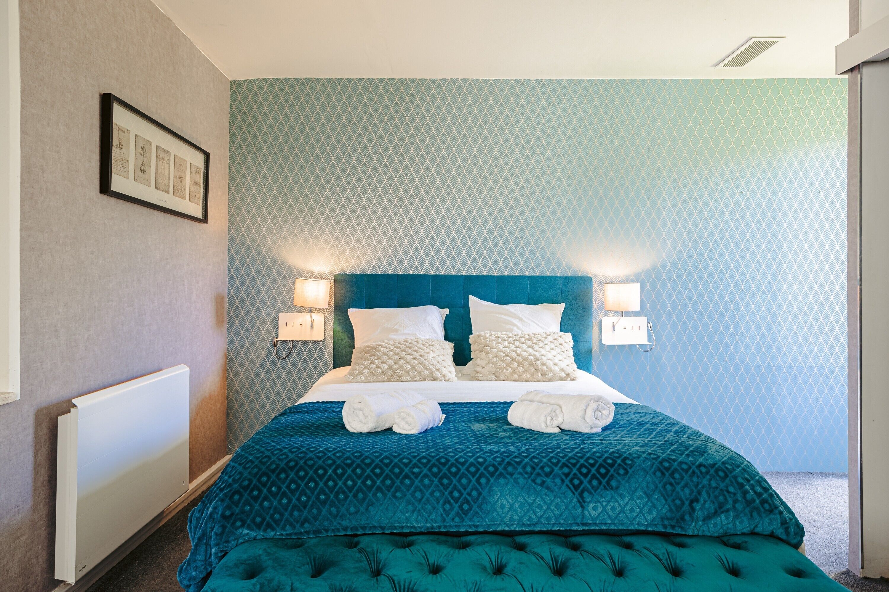 A hotel room with a bed, two wall-mounted lights, a framed picture, and a radiator.