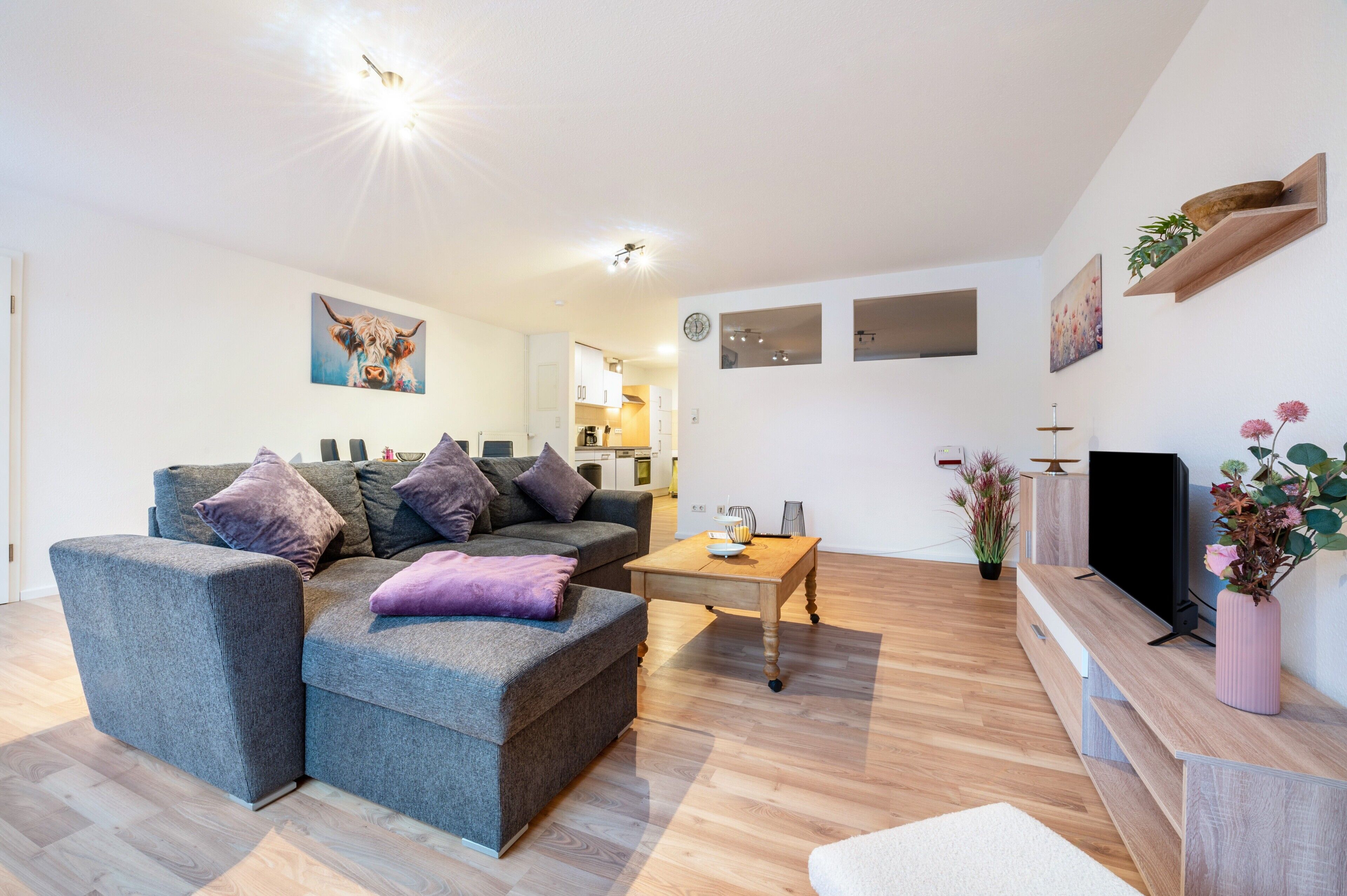 Ein modernes Wohnzimmer mit grauem Sofa, einem Holztisch, einem Flachbildfernseher und einer kleinen Pflanze.