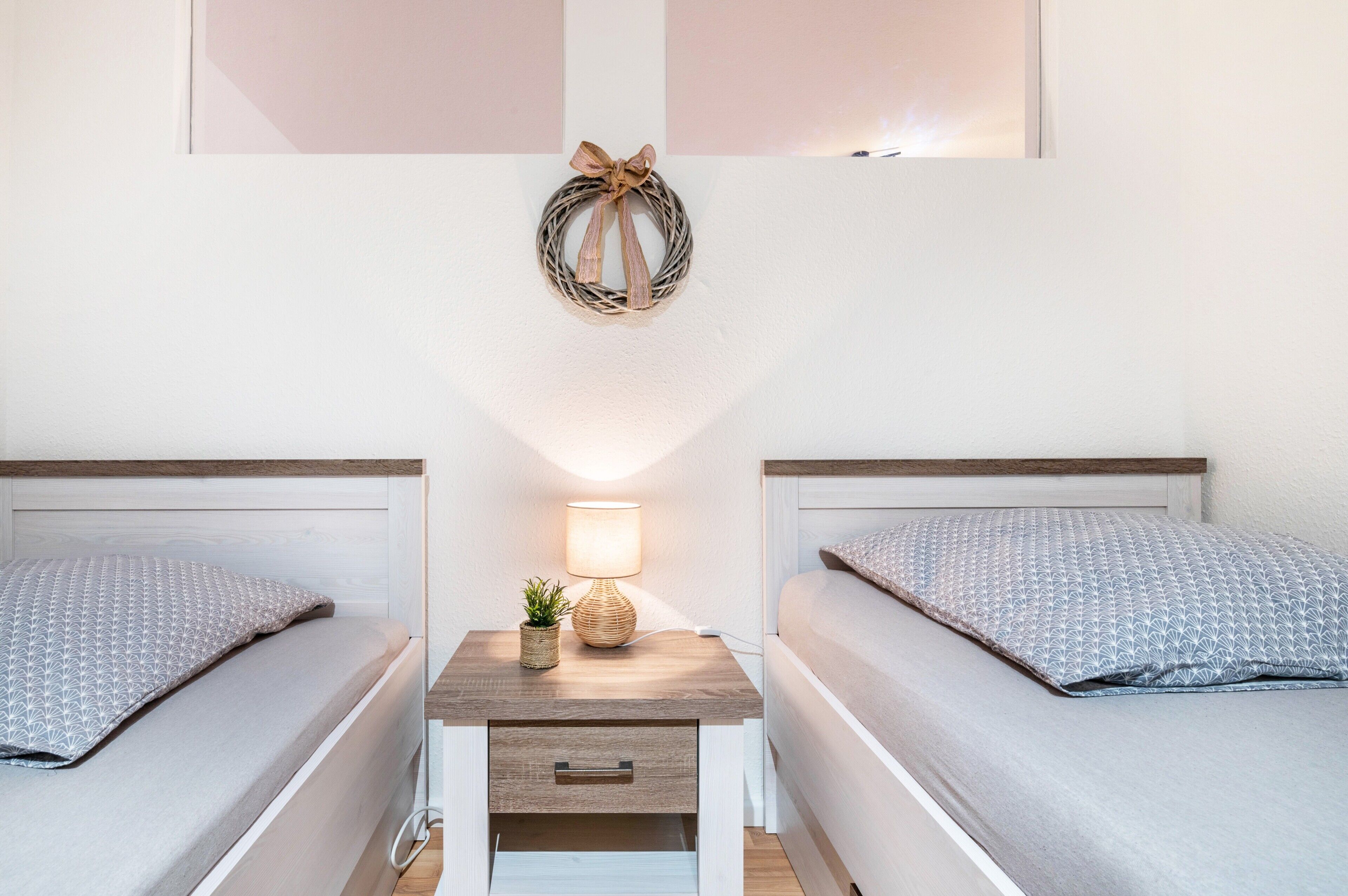 Ein Doppelzimmer mit zwei Betten, einem Nachttisch mit Lampe, einer kleinen Pflanze und einem Wandkranz.