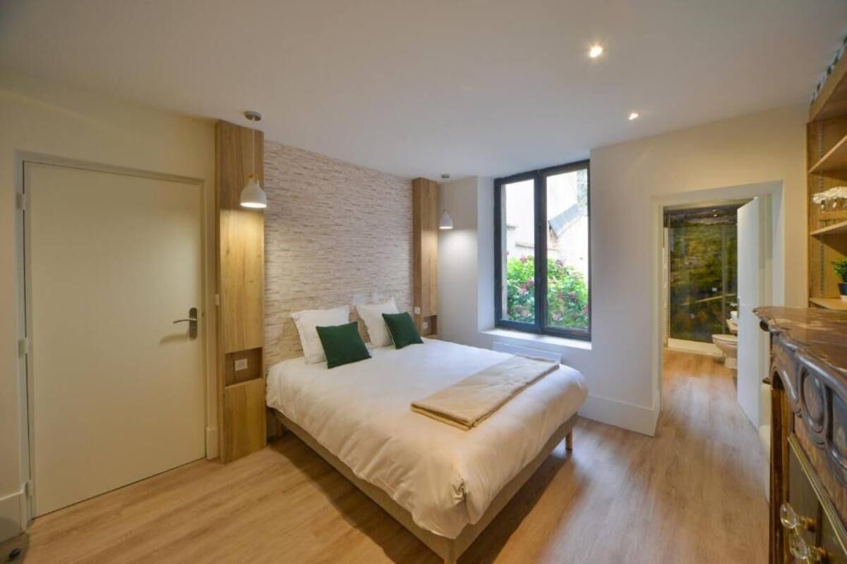 A bedroom with a large bed, a wooden wardrobe, a window with a view of greenery, and a stone wall.