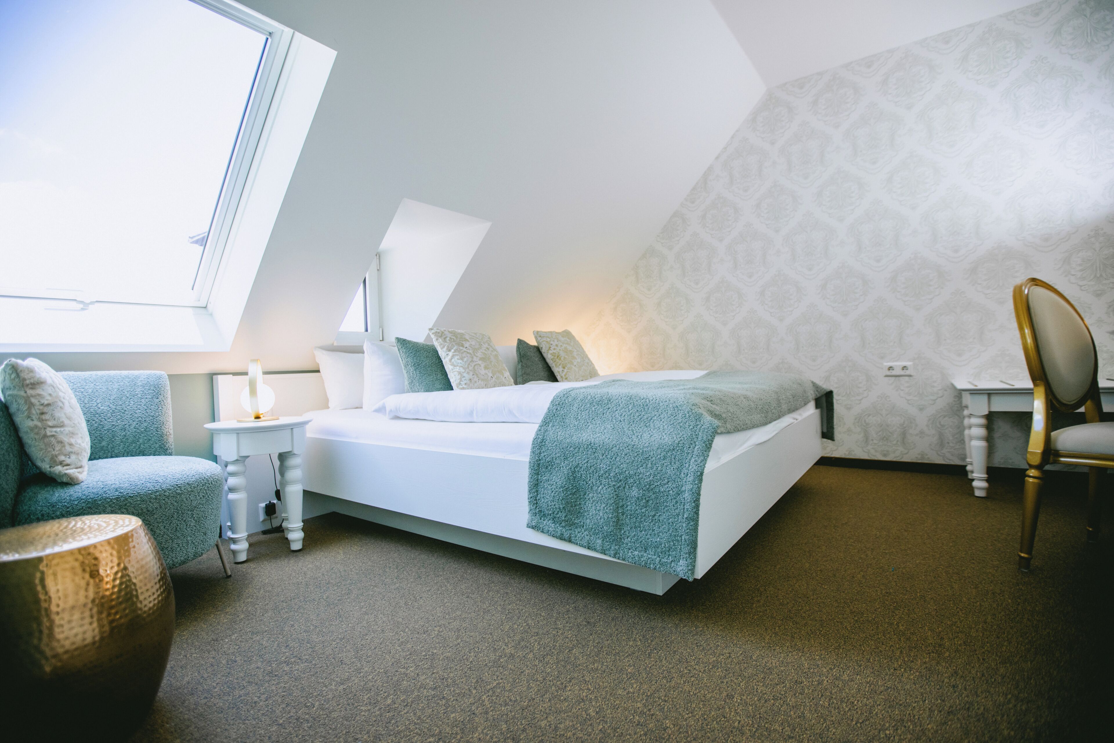 Ein Schlafzimmer mit schrägen Wänden, einem Bett mit weißem Kopfteil, einem Nachttisch mit Lampe, einem Sessel und einer Wand mit Muster.