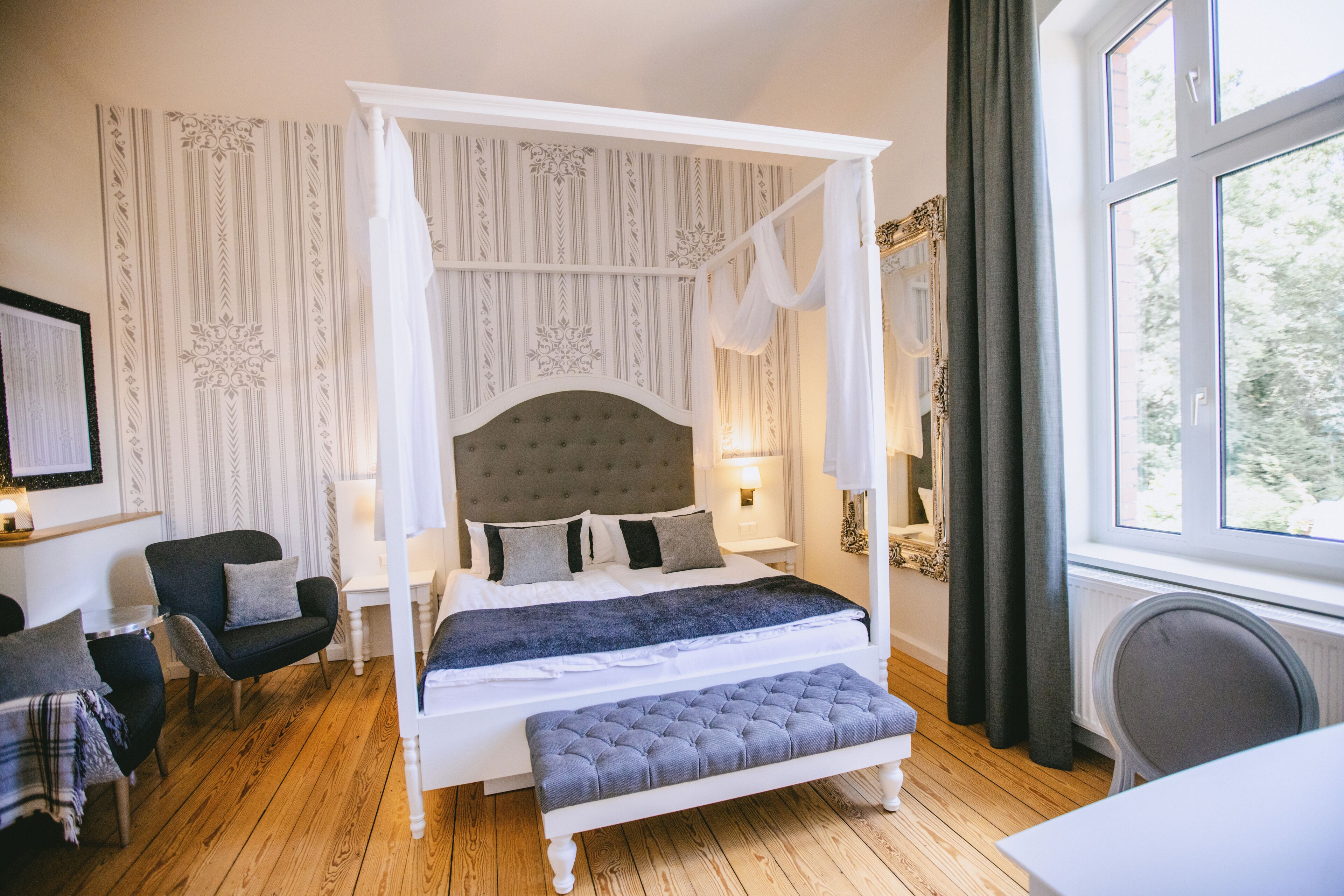 A four-poster canopy bed with a tufted headboard, a bench at the foot of the bed, and a view of the outdoors through a window.
