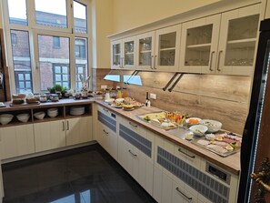 A modern kitchen with a breakfast buffet, wooden cabinets, and a large window overlooking a brick building.