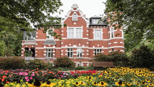 A large brick building with a symmetrical facade, a central entrance, and a surrounding garden with various flowers. - Johanniter Hotel Wittenberg (Lutherstadt Wittenberg)