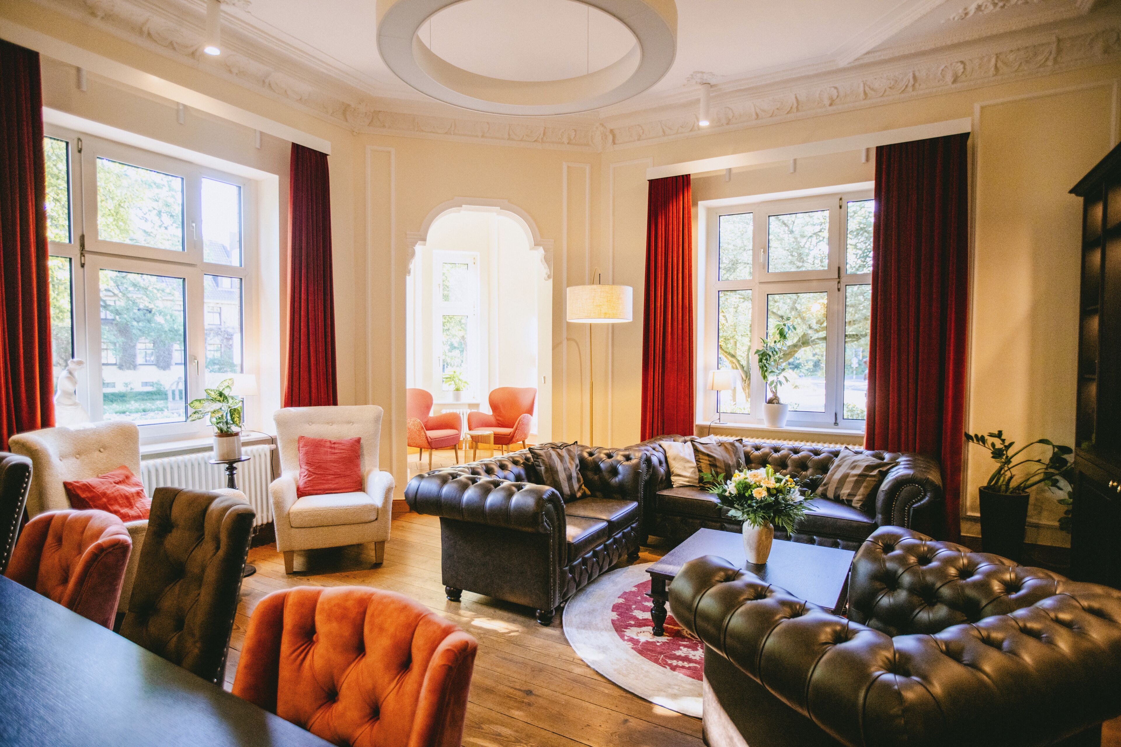 A spacious living room with a large sectional sofa, a round coffee table, and a fireplace. There are multiple windows providing natural light and a view of the outdoors.
