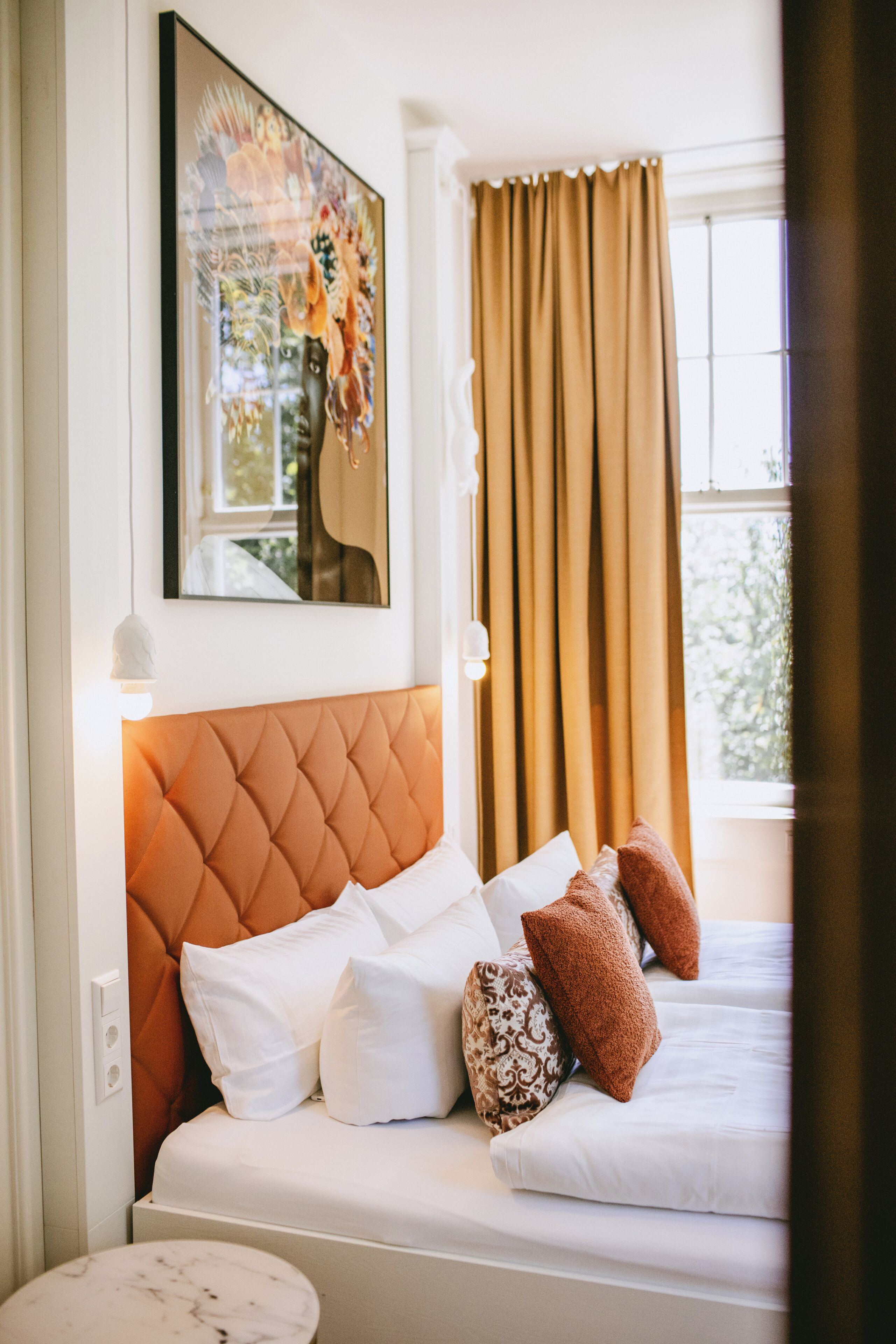 Hotelkamer met een bed, kussens, een hoofdeinde, een schilderij, een raam met gordijnen en een wandlamp.