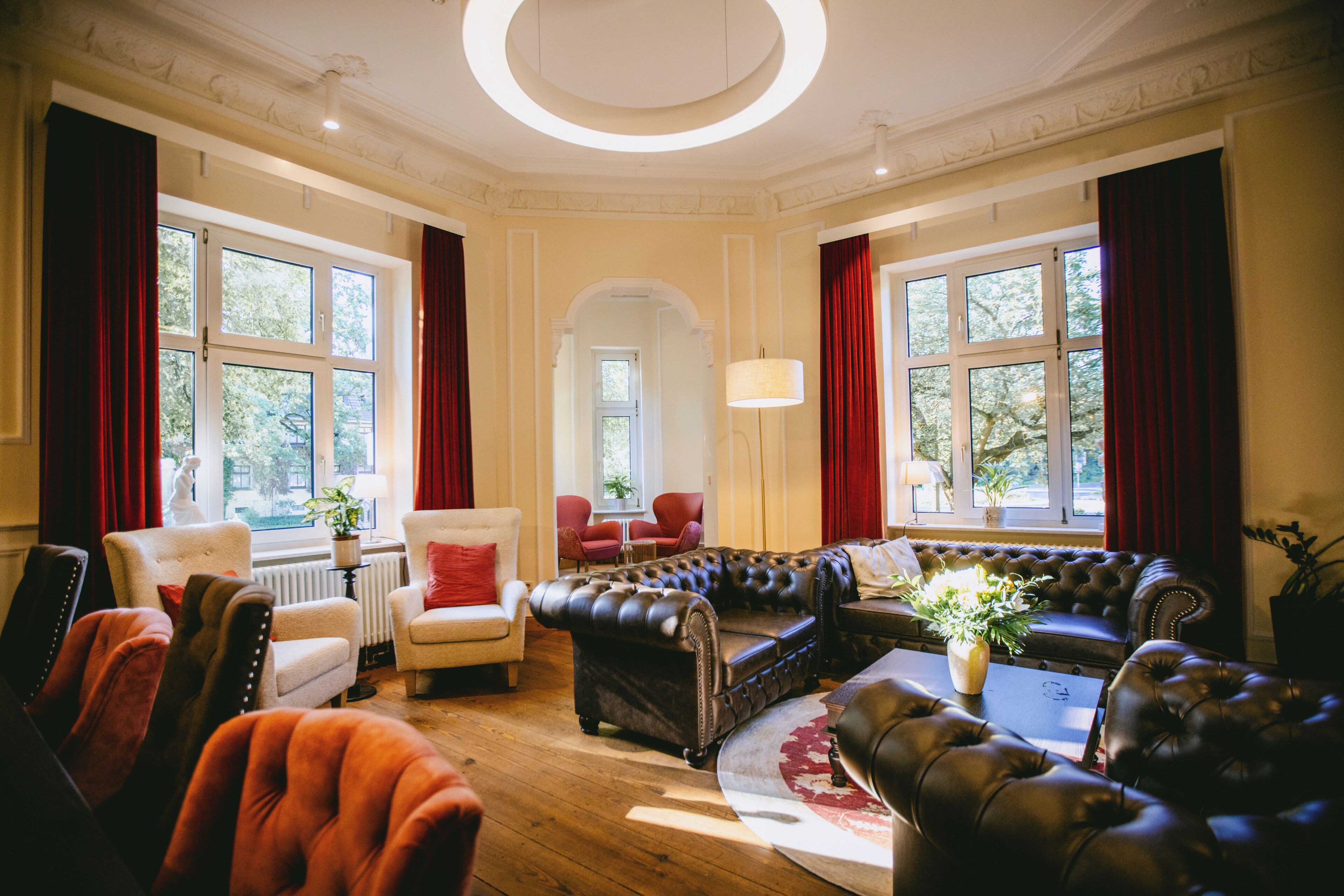 Amplia sala de estar con un conjunto de sofá de cuero, un sillón rojo y un sillón blanco. Hay ventanas grandes con cortinas rojas, una araña y una planta en el alféizar.