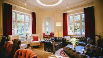 Amplia sala de estar con un conjunto de sofá de cuero, un sillón rojo y un sillón blanco. Hay ventanas grandes con cortinas rojas, una araña y una planta en el alféizar.