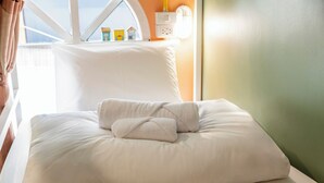Un lit bien fait, avec du linge de lit blanc et une serviette pliée, une fenêtre donnant sur des maisons et une prise électrique au mur.
