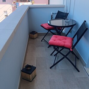 Ein kleiner Balkon mit zwei schwarzen Stühlen und einem Tisch, einer Mülltonne und einem Fenster mit Blick auf Gebäude.