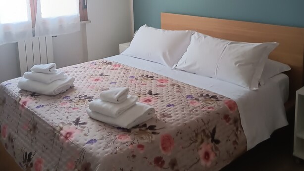 A neatly made bed with white linens and floral-patterned bedding, a painting of flowers on the wall, and a window with sheer curtains. - ‘Tosca 5 L.T.I.’ Apartment with Balcony and Air Conditioning (Pisa)
