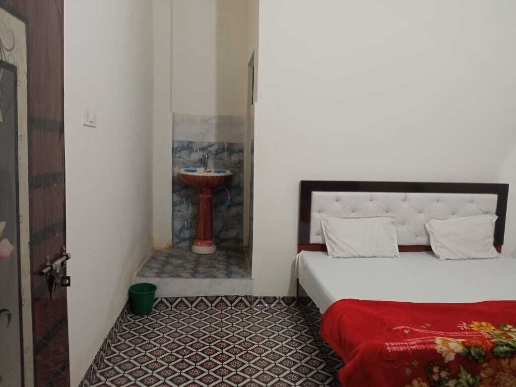 Une petite pièce, de taille modeste, comprenant un lit, une couverture rouge, un seau vert et un lavabo dans un coin.