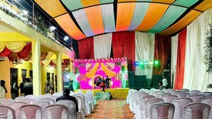Ein Saal mit Reihen von mit weißem Stoff bezogenen Stühlen, einer Bühne mit bunten Vorhängen und einem gemusterten Boden.