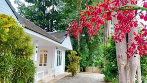 A white house with a gravel driveway, surrounded by lush greenery and a tree with red flowers. - Silpasom ARTstay Suratthani (Phunphin)